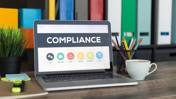 A laptop displaying the word "Compliance" on the screen. On the desk beside it are a cup of coffee, sticky notes, and a highlighter.